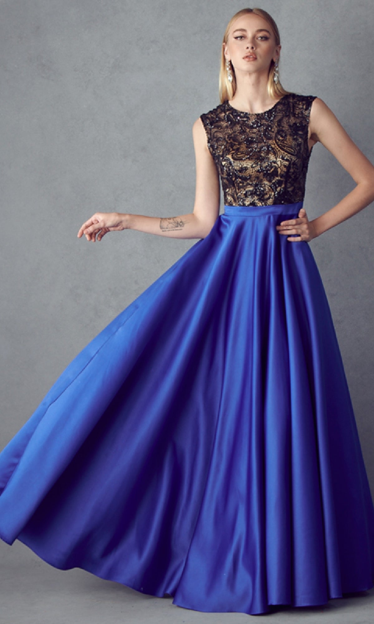 Woman wearing the Juliet 661 Cut-Out Long A-Line Prom Dress in Royal/Black, featuring an embellished bodice and sleeveless, floor-length satin skirt, standing against a plain gray background.